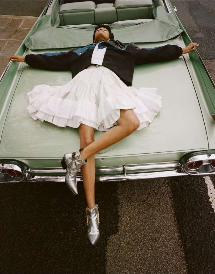 Woman lies on a light green convertible in a white ruffled mini dress and silver cowboy boots on a dark asphalt road outdoors.