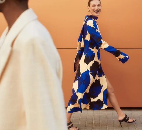 Woman in blue and cream floral print dress walking outdoors past a warm orange wall, photographed by Agata Pospieszynska.