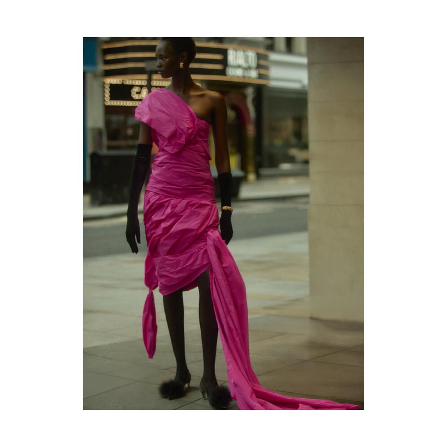 A woman walks in a stylish pink draped dress, black gloves, and heels on a city sidewalk with a theater in the background.
