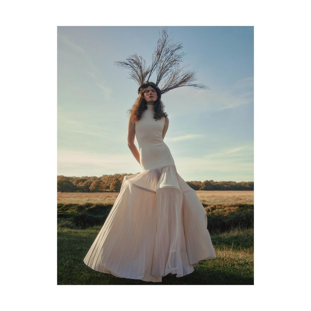 Fashion shot of a young adult woman in a white dress with a feather headpiece, standing outdoors in a grassy field under a blue sky.