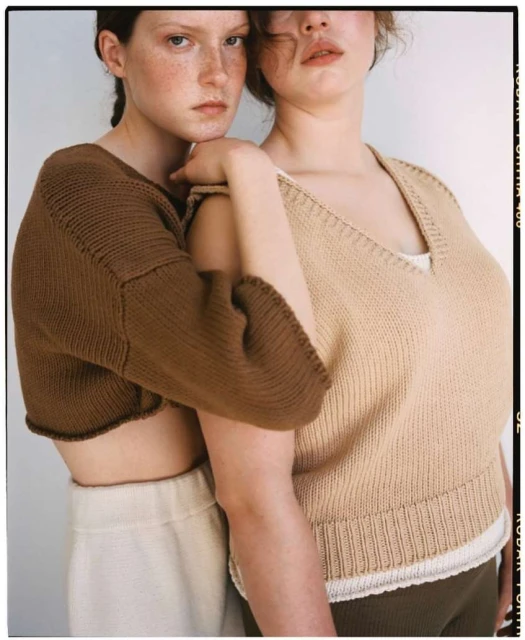 Two fair-skinned young women pose in knit tops and neutral pants against a plain background, with film border and soft lighting.