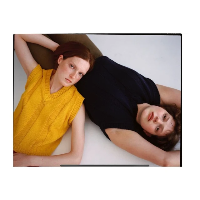 Two women lie side-by-side indoors wearing knit sweater vests, one yellow and one black, in a fashion editorial-style studio shot.