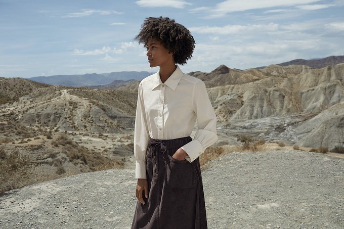 Woman in long-sleeved white shirt and dark skirt stands in arid landscape with rocky hills and blue sky with light clouds.