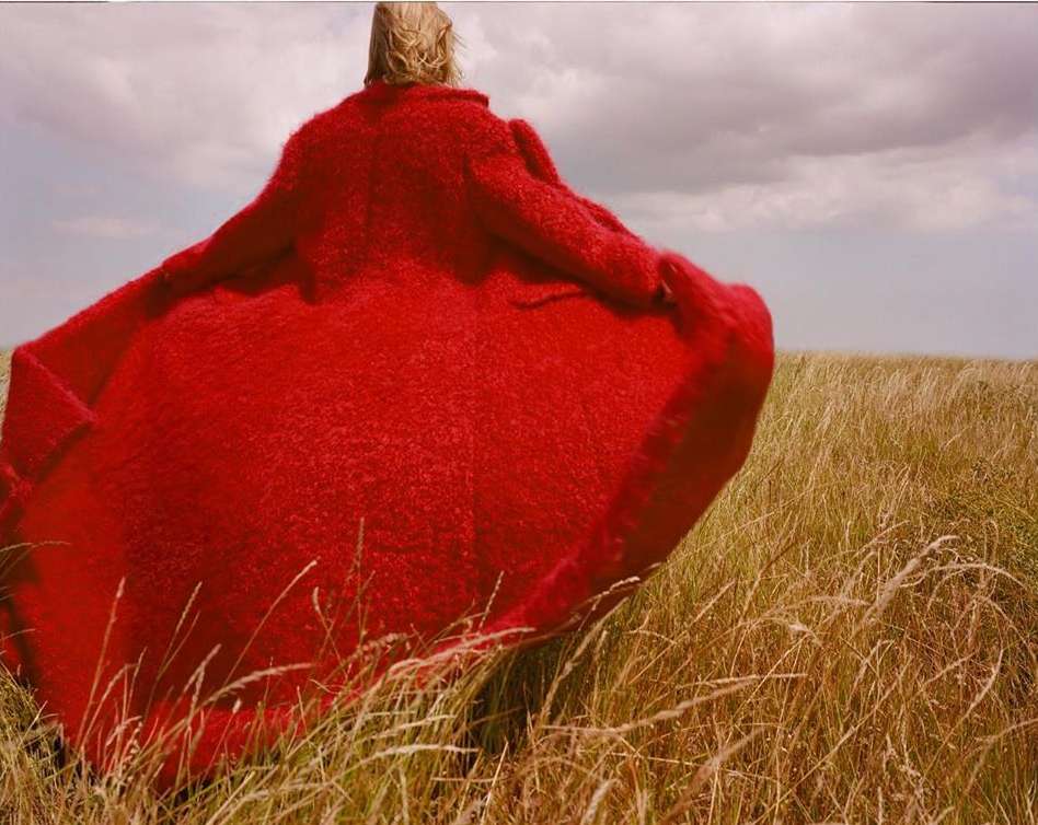 Back view of a blond adult woman wearing a large red coat walking through a field of tall brown grass with a cloudy sky background.