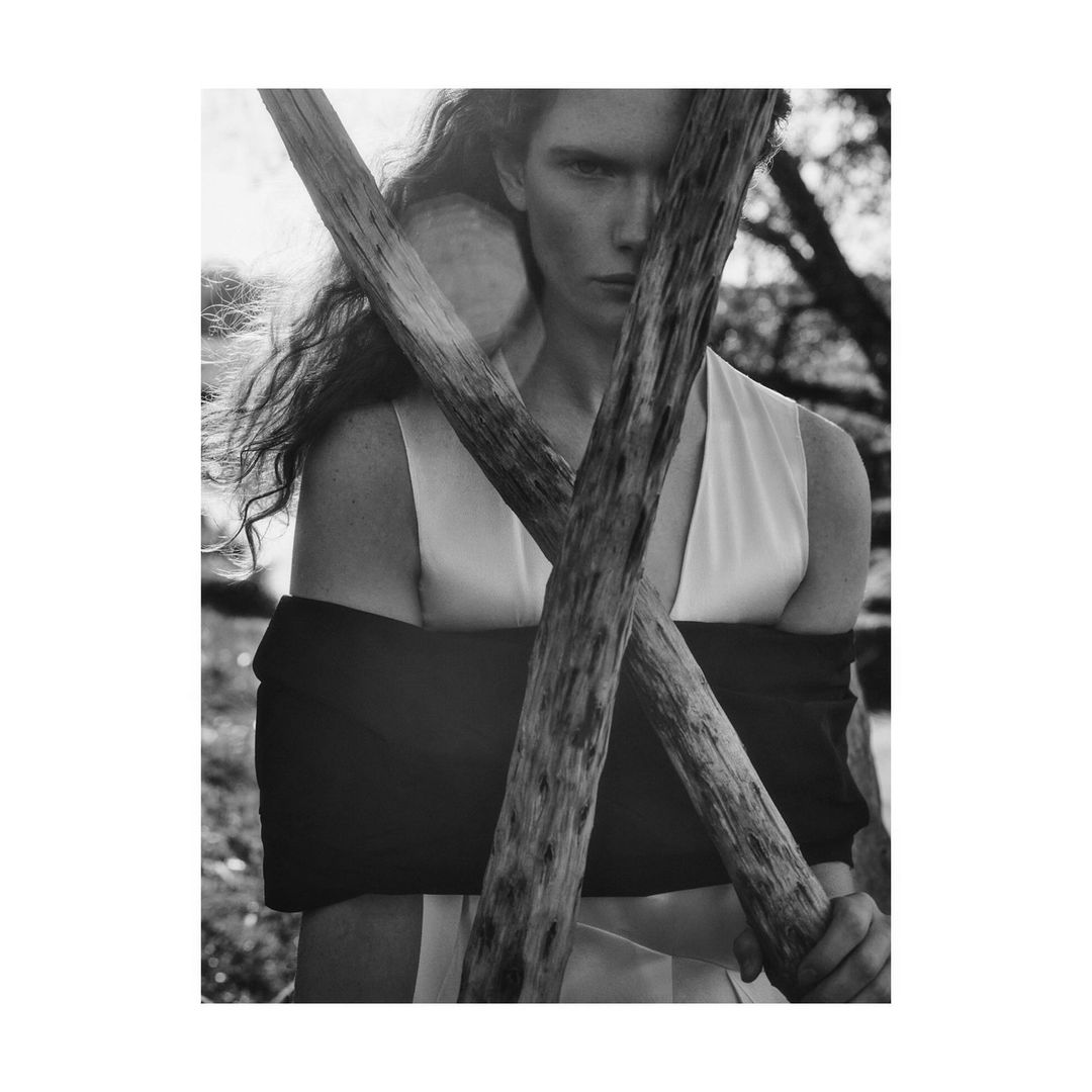 Monochrome outdoor portrait of a woman in sleeveless top, holding a wooden branch, with soft sunlight and trees in the background.