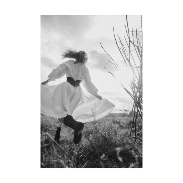 Black and white outdoor shot of a female in a white dress, moving through a grassy field with sparse branches reaching up to the sky.