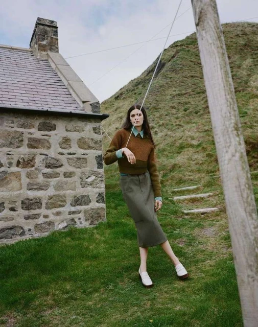 A woman in a brown sweater and skirt stands in a rural setting by a stone house and grassy hill beneath a cloudy sky.