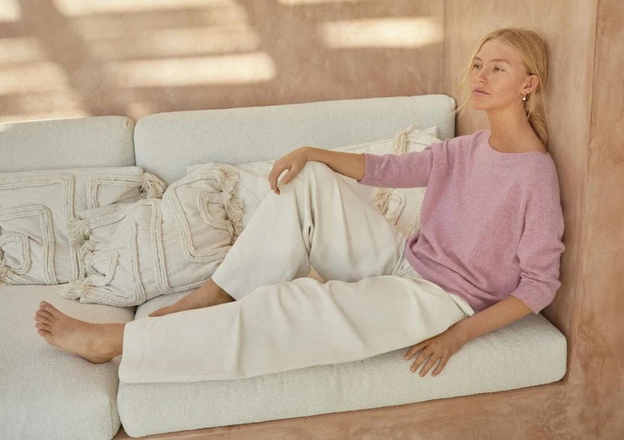 Woman in a pink sweater and white pants sits on a white textured couch with decorative pillows inside a room with light peach walls.