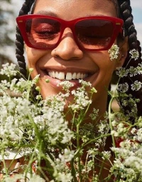 Close-up portrait of a smiling Black woman wearing red sunglasses, surrounded by white flowers.