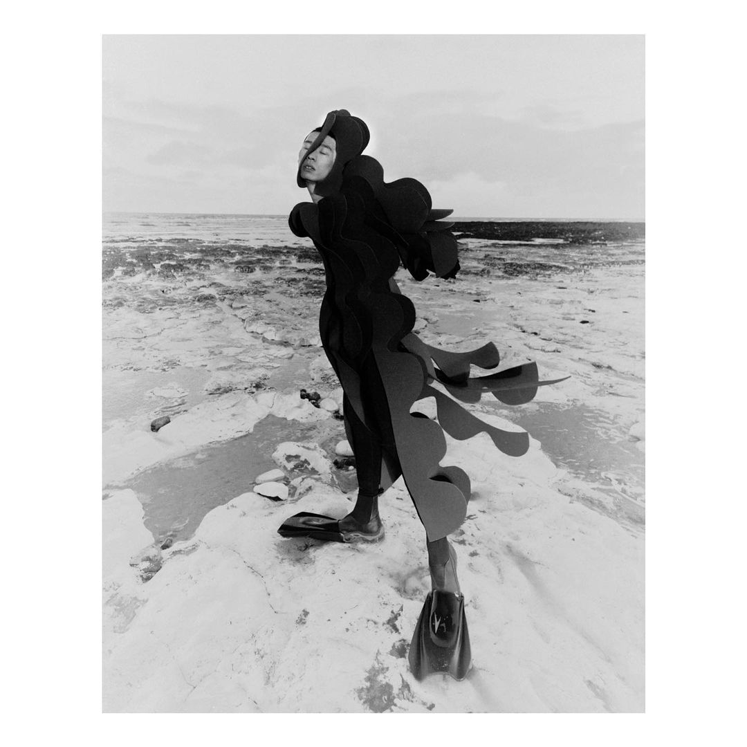 Monochrome image of a person in a black wetsuit and swim fins standing on a rocky beach, facing forward, near the ocean on an overcast day.