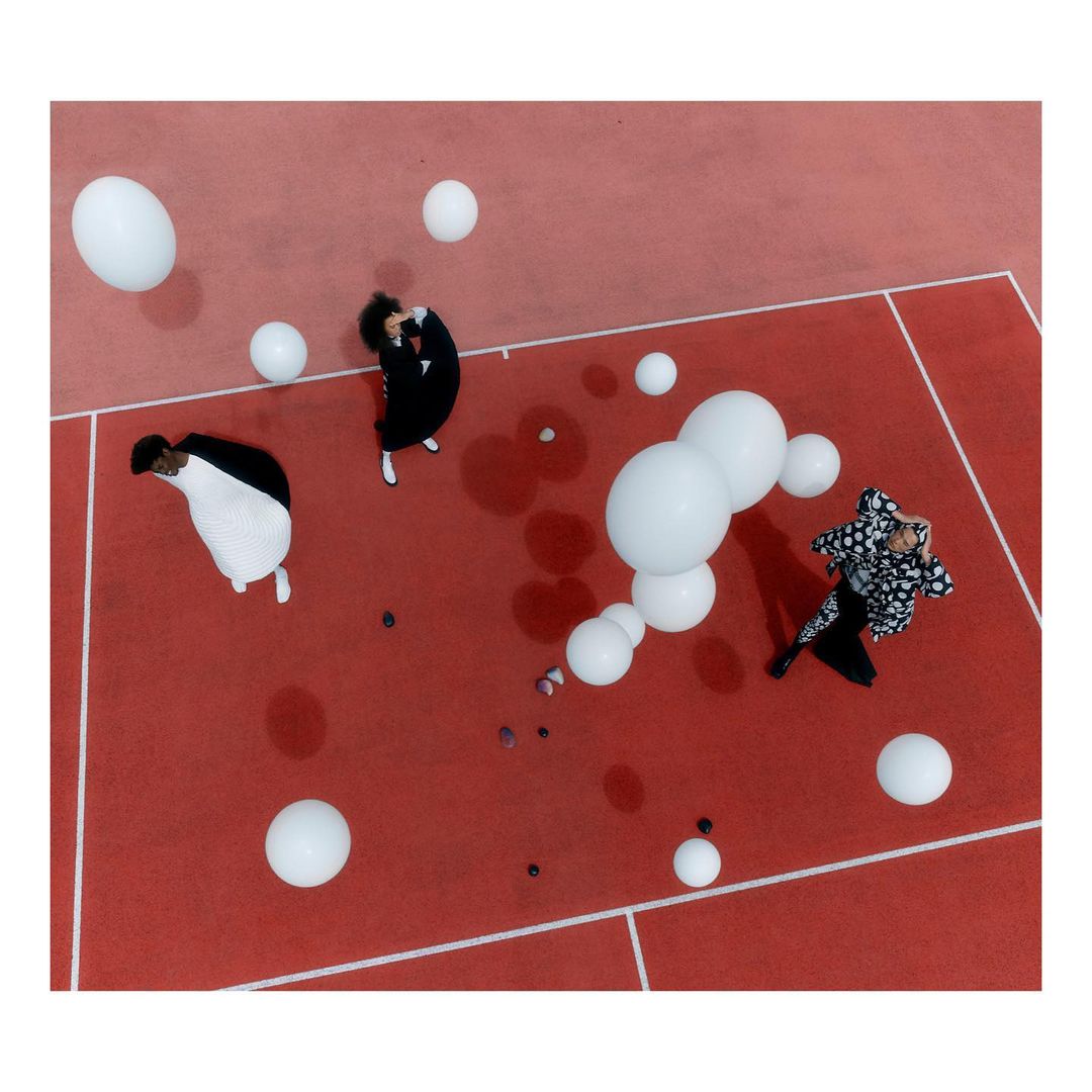 Aerial view of group of people in black and white costumes lying on red surface with scattered white spherical balloons overhead.