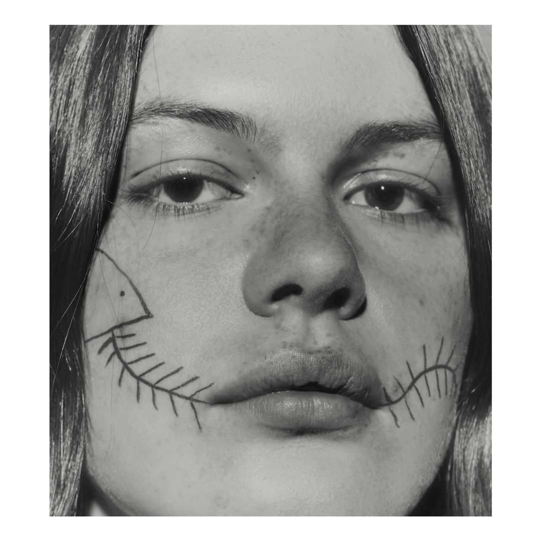Close-up black and white portrait of a young adult with drawing of a fish skeleton along their jawline in a studio setting.