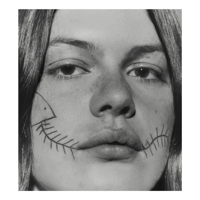 Close-up black and white portrait of a young adult with drawing of a fish skeleton along their jawline in a studio setting.