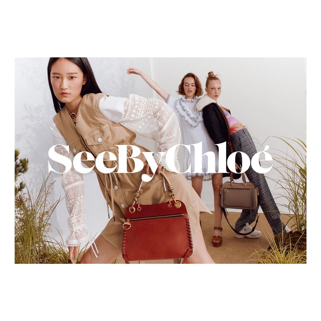 Fashion campaign featuring three female models in a studio setting. They're styled in coordinated outfits, carrying handbags, and posing dynamically.