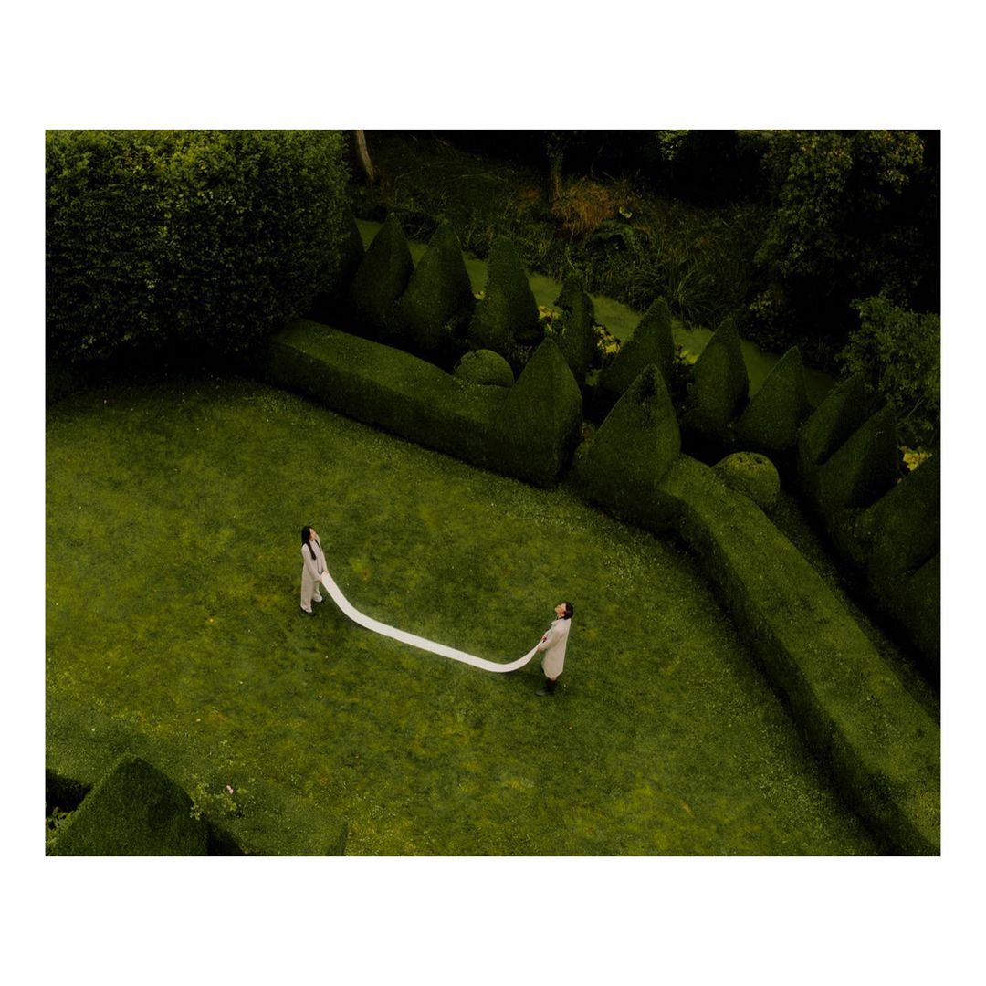 Two adult females in neutral coats hold a long white sheet in a green garden with trimmed hedges, captured in a high-angle view.