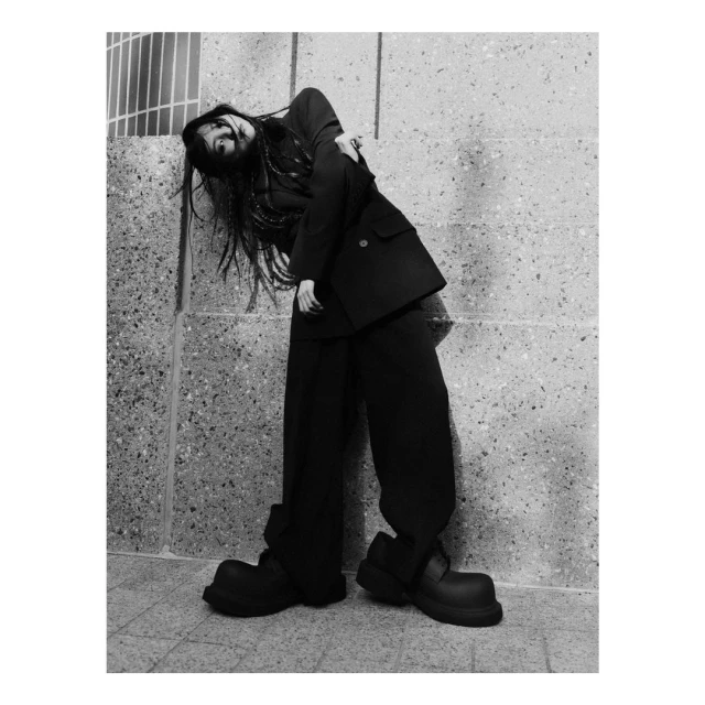 Monochrome fashion shot of a woman in a black suit and large black shoes, posing against a textured concrete wall in natural light.