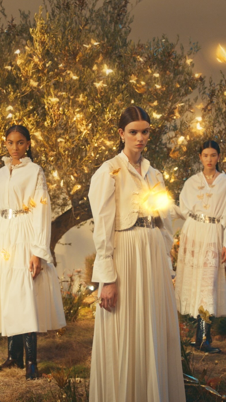 Three women in long white dresses pose outdoors surrounded by foliage and illuminated butterflies in a fashion editorial style image.