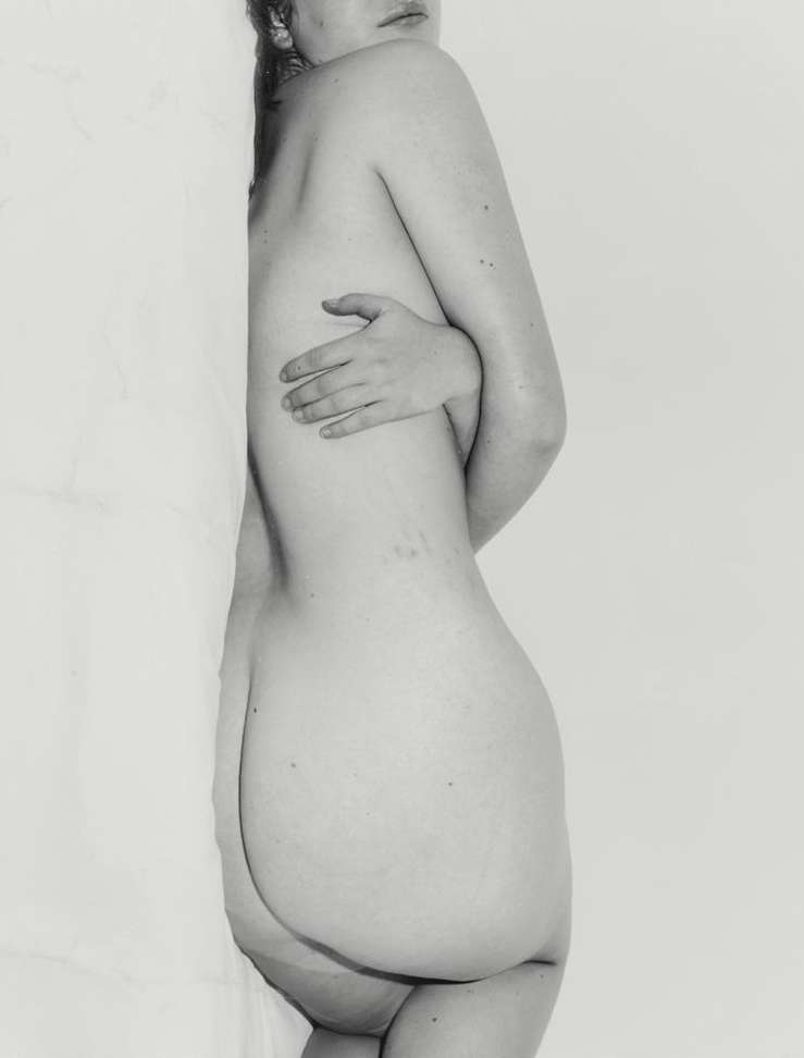 Monochrome portrait of a nude woman seen from the side, leaning against a white studio wall