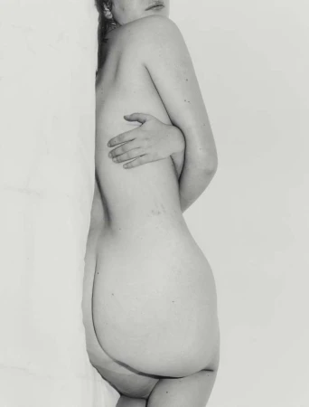 Monochrome portrait of a nude woman seen from the side, leaning against a white studio wall
