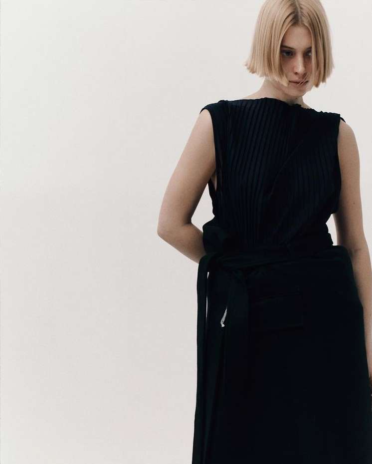 Woman wearing a dark, sleeveless top and skirt with a wrap detail against a light background in a fashion-style photograph.