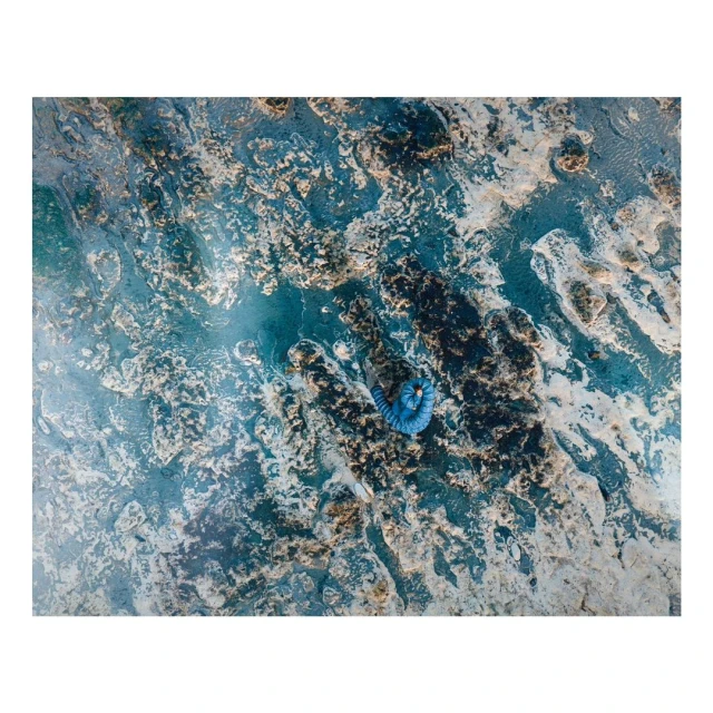 Overhead shot of a woman in blue wading in shallow blue water surrounded by rocky formations and sand, creating an abstract seascape.
