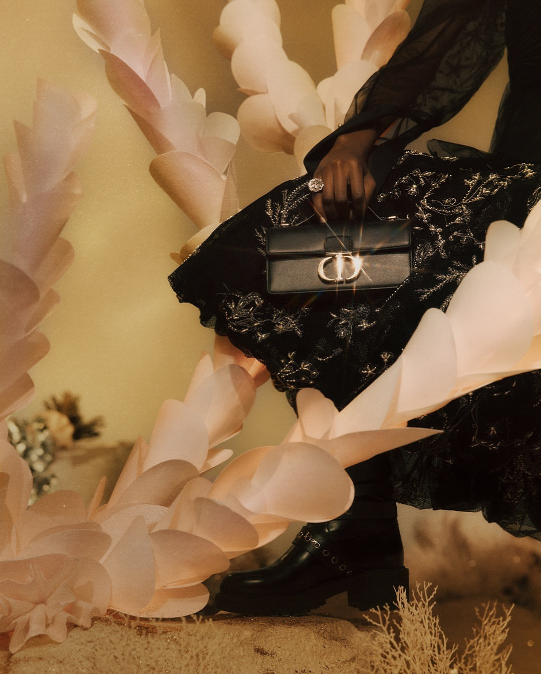Woman wearing a black dress and boots, holding a Dior purse with light pink decorative leaves and tan ground in a fashion setting.