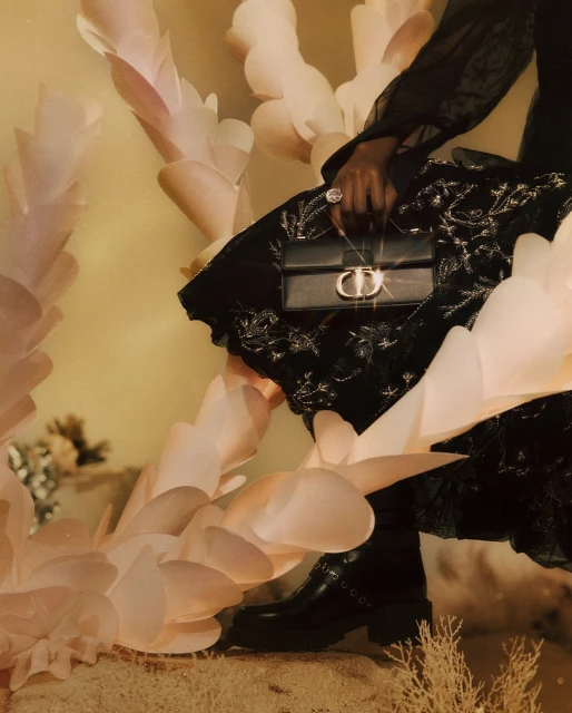 Woman wearing a black dress and boots, holding a Dior purse with light pink decorative leaves and tan ground in a fashion setting.