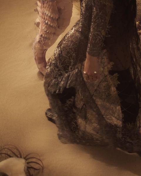 Close-up on a person in an ornate dark gown next to a large, pale sculptural element resembling feathers, possibly on a stage.