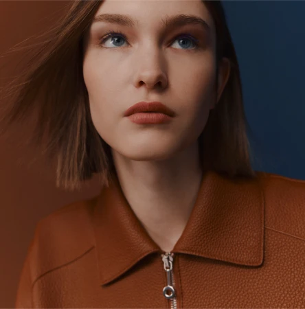 Close-up of a young woman with short brown hair and blue eyes, wearing a brown leather jacket, against a blue and brown background.