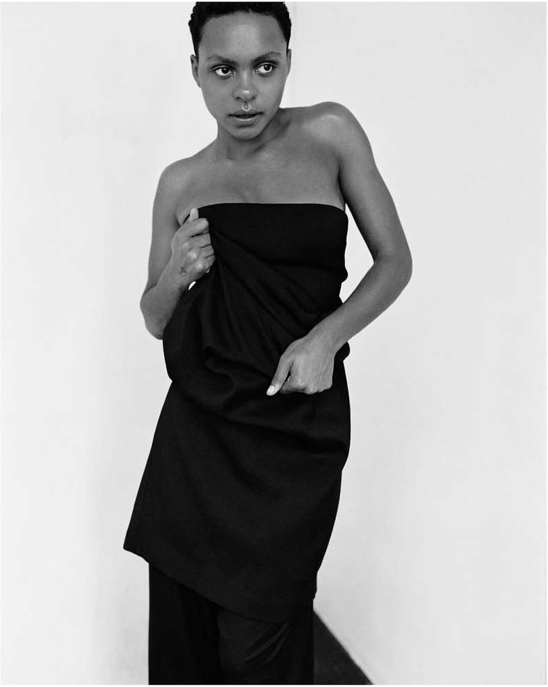Monochrome fashion portrait of a Black woman wrapped in draped fabric, gazing softly, against a plain white background in studio lighting.