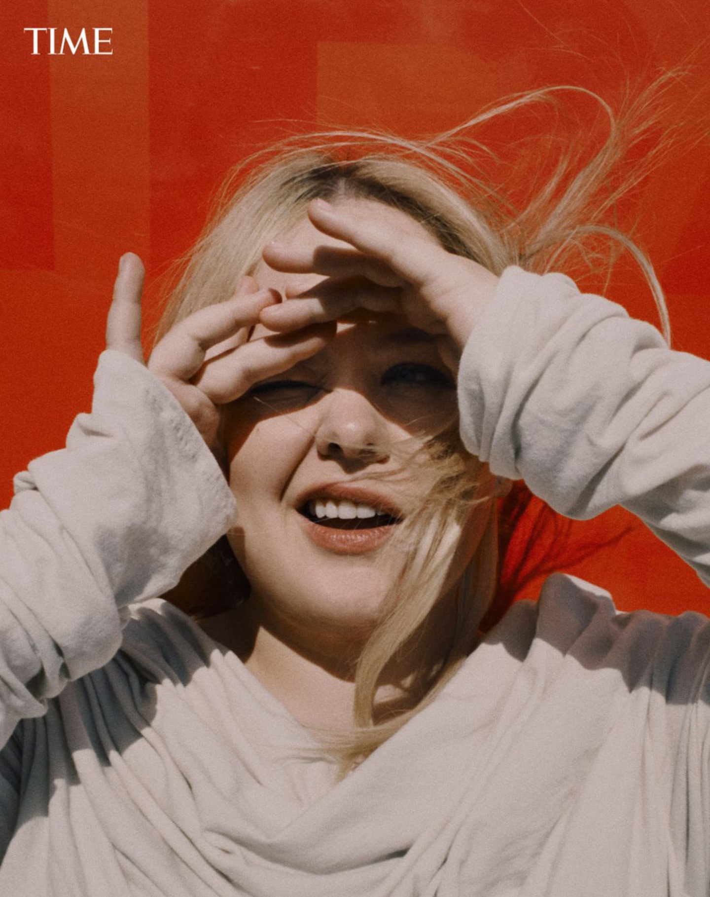 Portrait of smiling woman with blonde hair and fair skin wearing a neutral-toned top against an orange and red color block background.