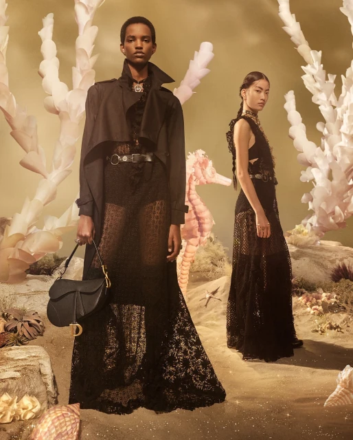 Two women pose in lace dresses and trench coats with a dark saddlebag against an oceanic backdrop with corals and sea shells.