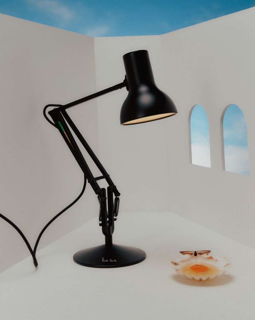 Black adjustable desk lamp on a white table in front of white walls with arched windows revealing a blue sky and seashell dish.