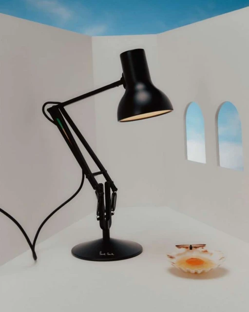 Black adjustable desk lamp on a white table in front of white walls with arched windows revealing a blue sky and seashell dish.