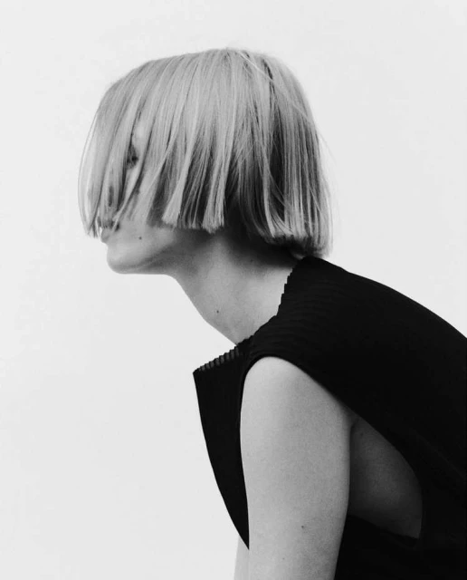 Black and white portrait of a person with a blonde bob hairstyle and a black sleeveless top against a neutral backdrop in a photo studio.