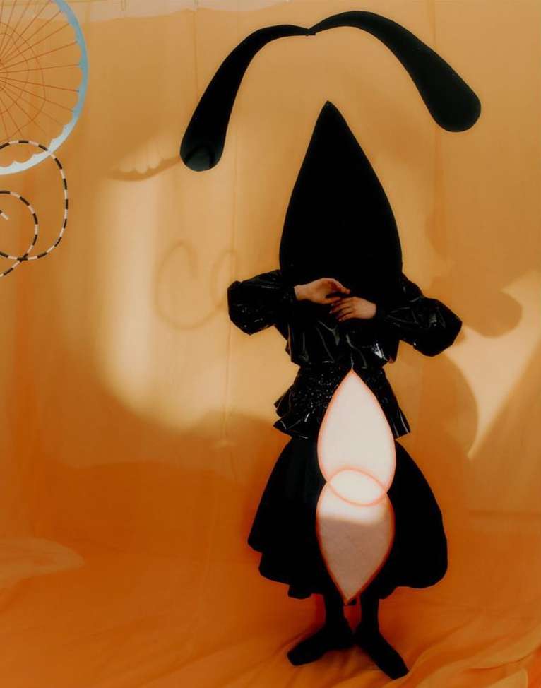 Person in black cone hat and glossy coat poses on peach background with bicycle wheel and dark abstract shapes, creative fashion shot.