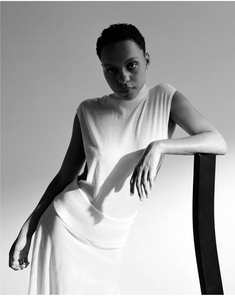 Black and white portrait of a woman with short hair wearing a light-colored sleeveless dress, leaning against a black chair.