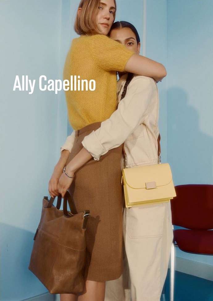 Two women pose in a fashion shot displaying bags; one woman wears a yellow sweater and the other is in a cream-colored jumpsuit.
