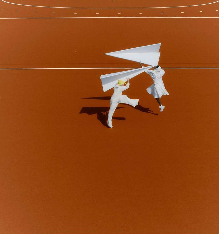Two people in white outfits hold up large paper airplanes on a terracotta-colored court with white lines, creating a playful scene.