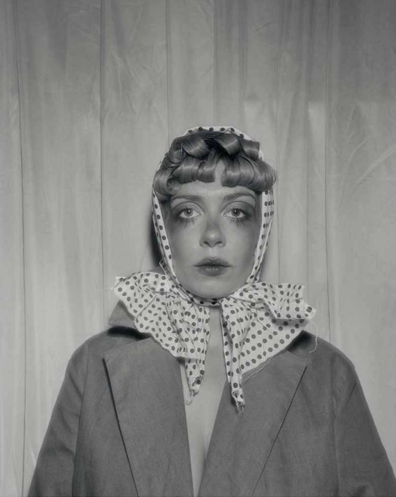 Monochrome portrait of an adult female with light skin, vintage hair, polka dot headscarf, and oversized jacket against a white curtain backdrop.