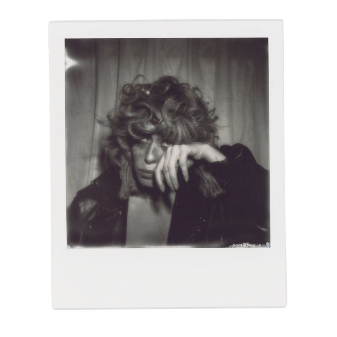 Black and white portrait of a person with curly hair wearing a jacket, hand to their face, against a curtain background.