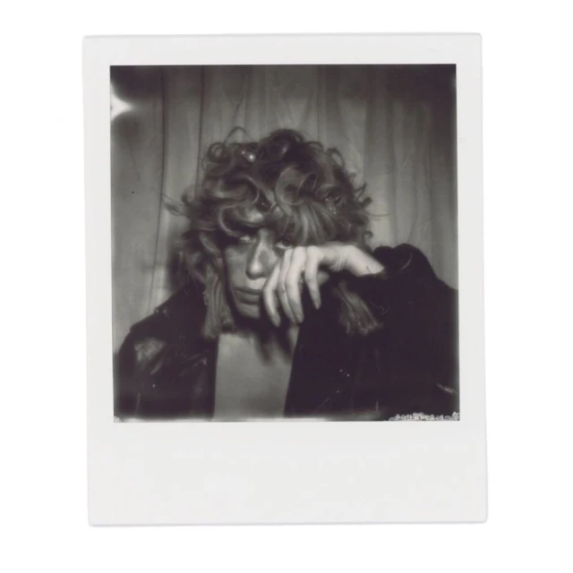 Black and white portrait of a person with curly hair wearing a jacket, hand to their face, against a curtain background.