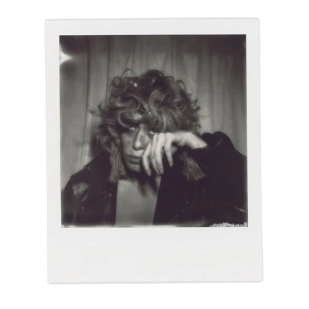 Black and white portrait of a person with curly hair wearing a jacket, hand to their face, against a curtain background.