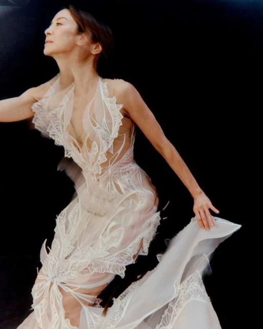 Elegant Asian woman in a flowing white gown poses against a dark background in a fashion editorial shot showcasing the gown's intricate design.