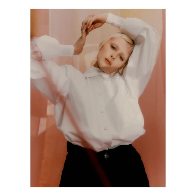 Portrait of a young woman with pale skin and blonde hair, wearing a white blouse, in a photo studio with colorful backdrop panels.