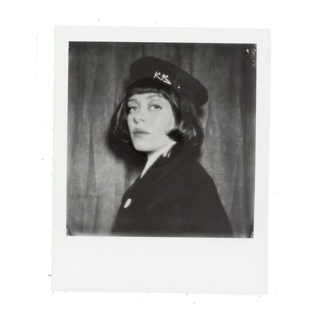 Monochrome portrait of an adult female in a dark coat and brimmed hat with visible text, against a neutral backdrop.