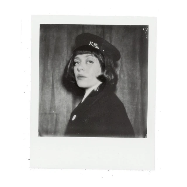 Monochrome portrait of an adult female in a dark coat and brimmed hat with visible text, against a neutral backdrop.