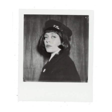 Monochrome portrait of an adult female in a dark coat and brimmed hat with visible text, against a neutral backdrop.