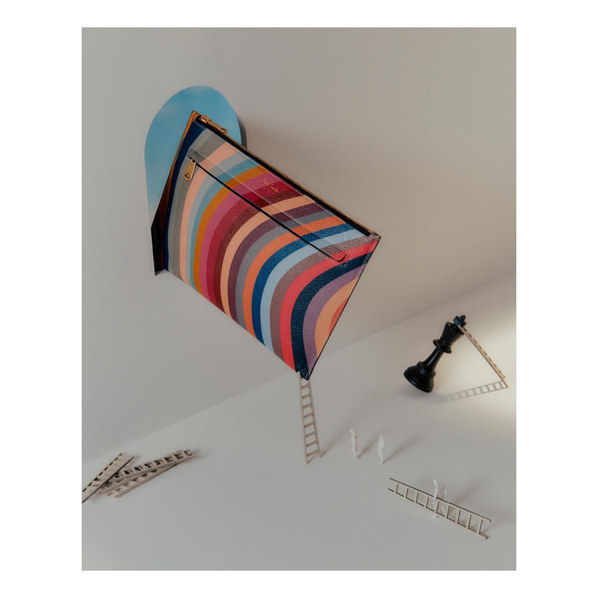 Conceptual product shot of a striped clutch purse attached to a blue shape on the wall, miniature ladders, and a chess piece.