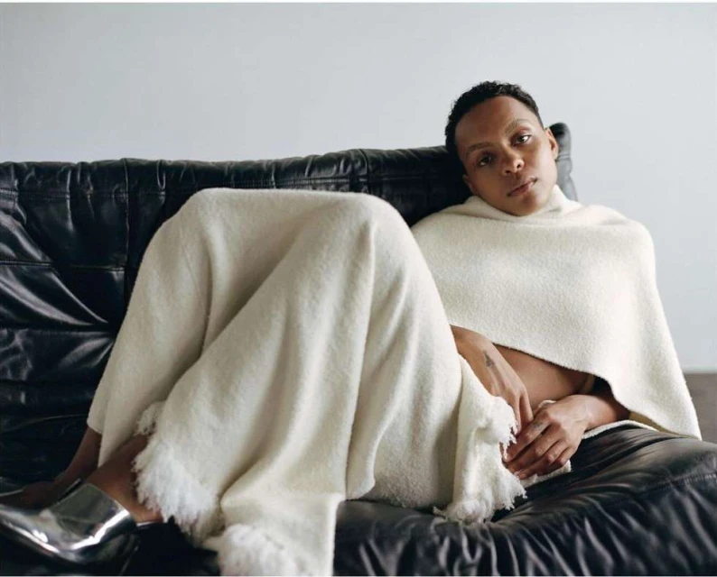 An adult with short hair sits on a black leather couch, draped in an off-white fringed blanket, with silver shoes and a neutral studio background.